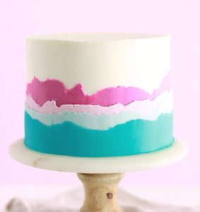 Round buttercream cake with white, pink and teal buttercream on a cake pedastal.