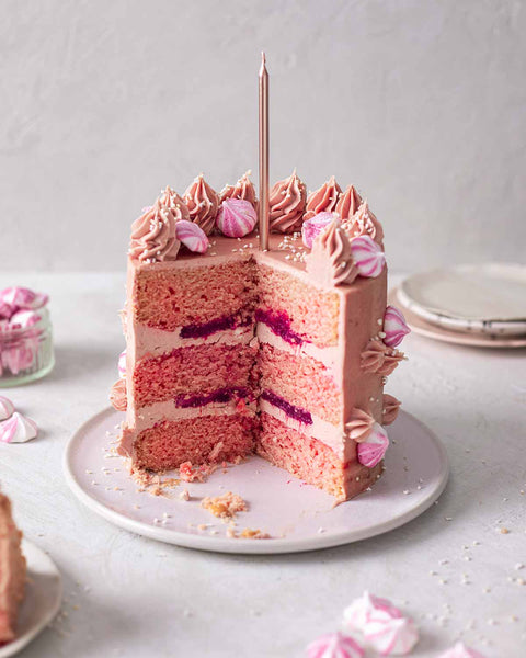 Three layer pink buttercream cake, sliced to see the jammy filling, topped with a tall gold candle. 