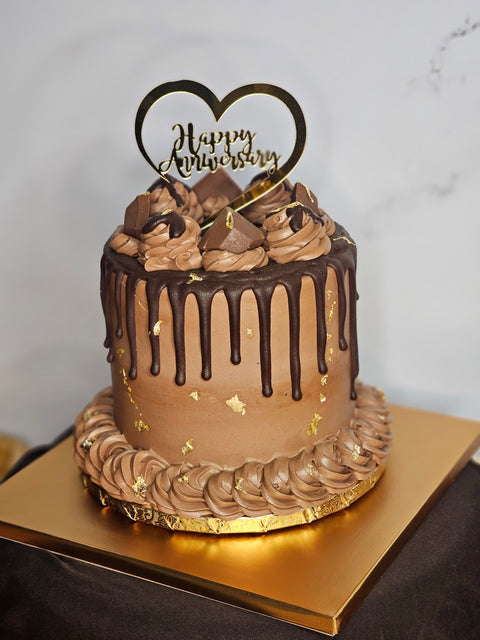 Close up of a chocolate buttercream cake decorated with chocolate buttercream, chocolate candies, gold flakes, and a Happy Anniversary plaque,  sitting on a gold cake board.