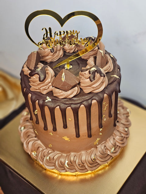 Close up, top angle  of a chocolate cake decorated with chocolate buttercream, chocolate candies, gold flakes, and a Happy Anniversary plaque,  sitting on a gold cake board.