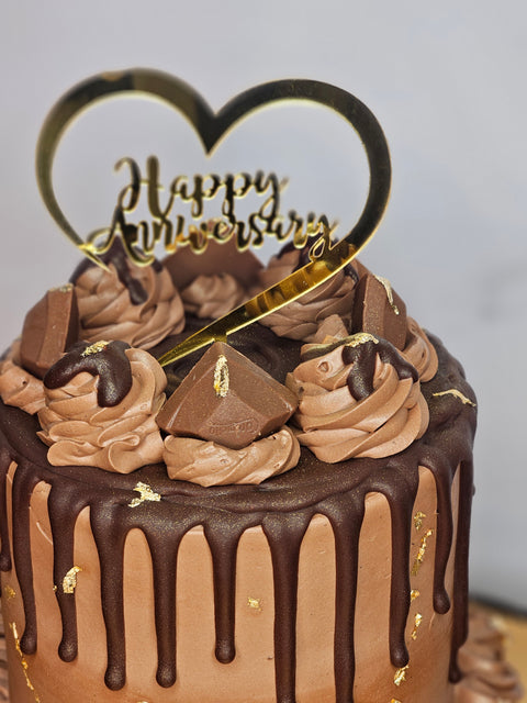 Super close up of the top of a chocolate buttercream cake decorated with chocolate buttercream, chocolate candies, gold flakes, and a Happy Anniversary plaque,  sitting on a gold cake board.