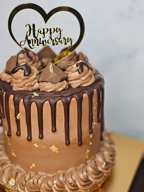 Close up of a chocolate buttercream cake decorated with chocolate buttercream, chocolate candies, gold flakes, and a Happy Anniversary plaque,  sitting on a gold cake board.