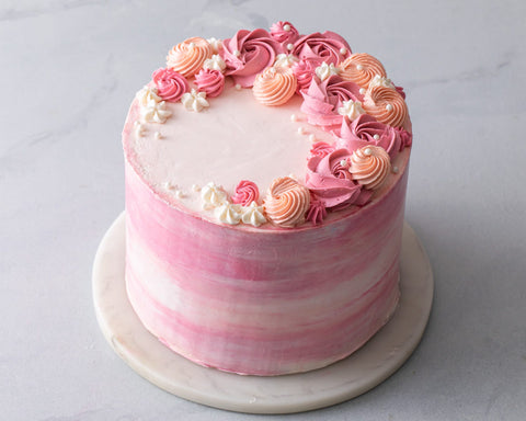 Pink watercolor buttercream on a white cake plate, decorated with buttercream rosettes.