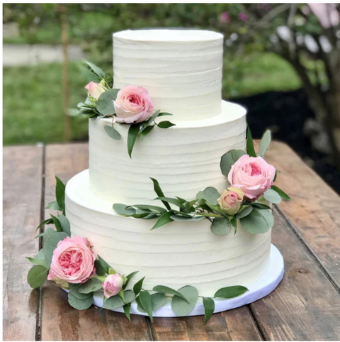 Three tiered cake decorated in white buttercrea, with pink flowers and green leaves, stting on a table.