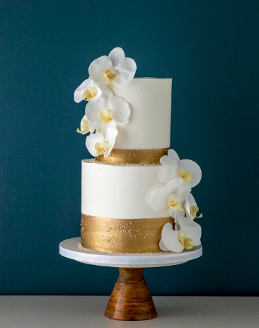 White buttercream cake doecorated with gold trim, and white flowers. With a teal background.