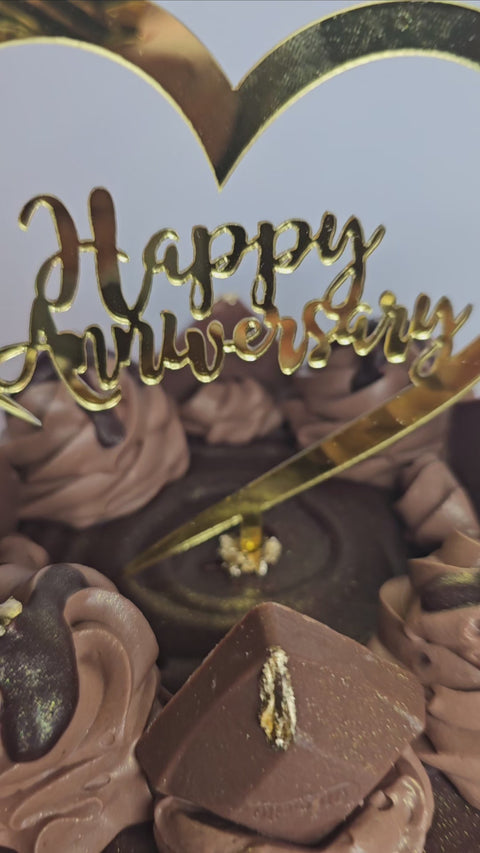 Close up of a chocolate cake decorated with chocolate buttercream, chocolate candies, gold flakes, and a Happy Anniversary plaque,  sitting on a gold cake board.